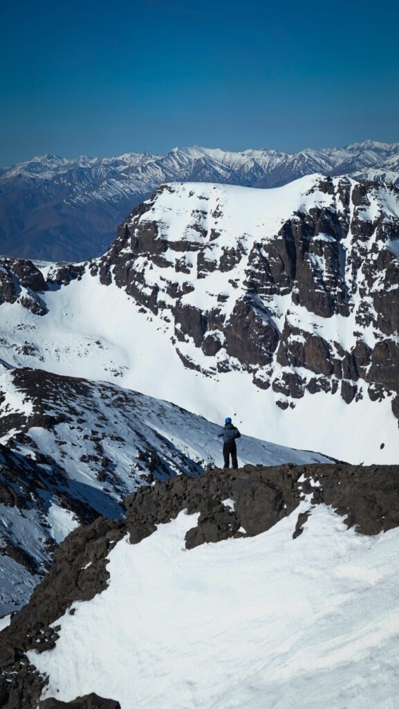 A lone explorer stands on a snow-covered peak in Morocco, surrounded by majestic mountainous terrain.