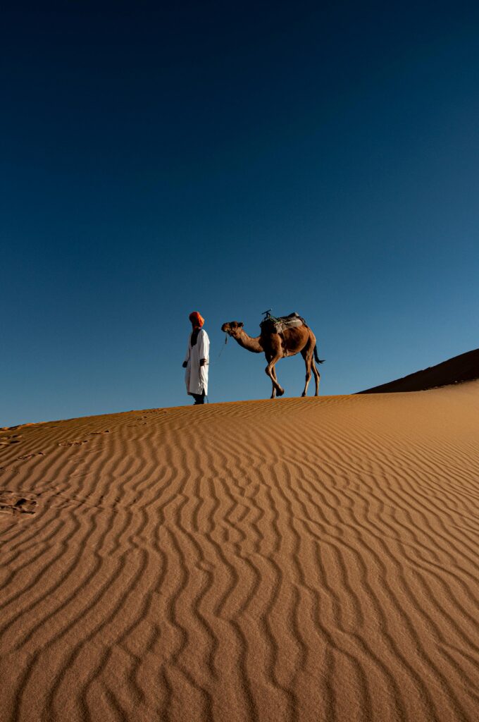 A man with a camel walks across a vast desert with beautiful ripple patterns under a clear blue sky.