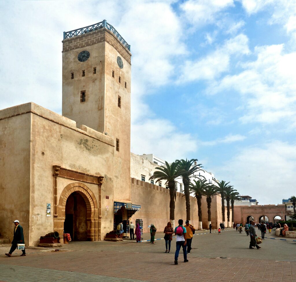 A scenic view of the historic city walls and clock tower in Essaouira, Morocco, with people walking by.
